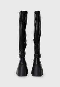Black knee-high boots made of faux leather with a flat platform sole, side zippers, and a buckle accent on the ankle.