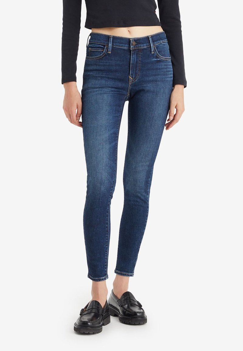 Personne portant un jean skinny bleu foncé, des mocassins noirs et un crop top noir à manches longues, debout devant un fond blanc uni.