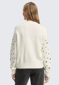 Biały sweter z teksturowanym wykończeniem, z czarnymi sercami i kropkami na rękawach. Żebrowane mankiety i dół zapewniające dopasowanie.