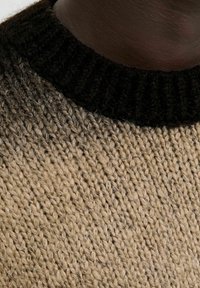 Nahaufnahme einer dunkelhäutigen Person, die einen dicken, beige gestrickten Pullover mit einem schwarzen Rippkragen trägt.