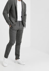 Šedý pletený cardigan s límcem, zapínání na knoflíky, v kombinaci se šedými kalhotami na míru a bílými teniskami. Jednoduchý, strukturovaný design.