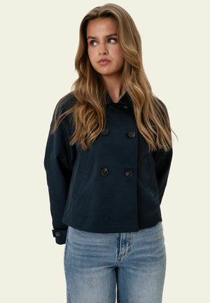 Donna con capelli lunghi e ondulati indossa una giacca doppiopetto blu navy e jeans azzurri, guarda verso destra su uno sfondo semplice.
