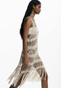 Pikkade mustade juustega naine kõndimas, seljas beežist lõngast kootud kleit frngiga valge triiksiti kohta ja sobivate kaelakee, valge tausta ees.