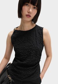 Czarny, strukturalny top bez rękawów o asymetrycznym dekolcie, z detalami ze złotego metalu na ramieniu oraz dopasowanym krojem.