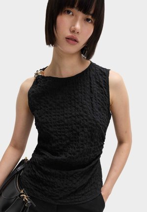 Zwart textuurloos mouwloos topje met een asymmetrische halslijn, voorzien van gouden hardware details op de schouder en een aansluitende pasvorm.