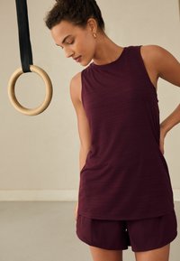 Top de sport burgundy cu un gât înalt, asortat cu pantaloni scurți. O inelastică de lemn pentru gimnastică atârnă deasupra, pe un fundal neutru.