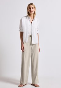 Chemise blanche à manches retroussées, associée à un pantalon beige à jambes larges. La tenue est complétée par une ceinture en tissu avec une boucle en métal.