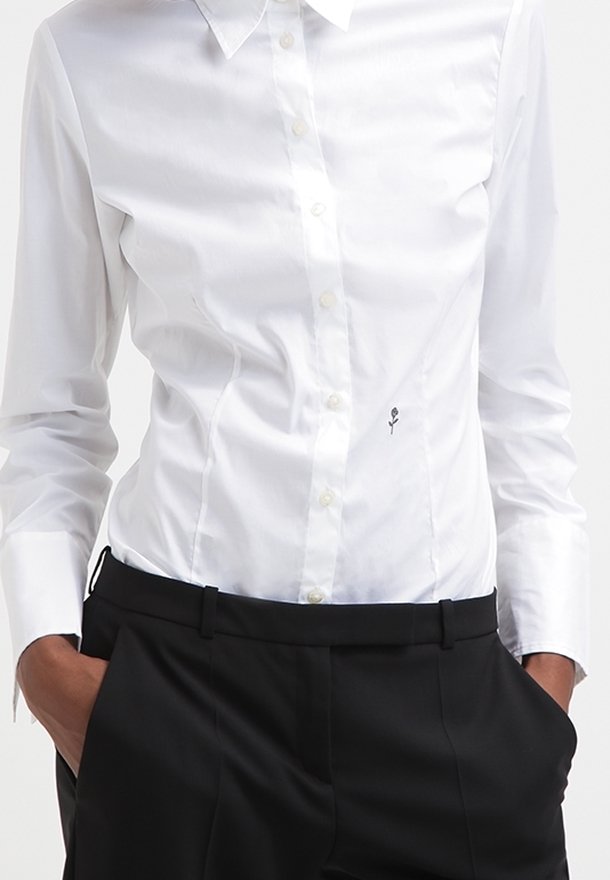 Camisa blanca de botón hecha de algodón suave, con un diseño ajustado y un pequeño bordado de rosa negra en el frente. Combinada con pantalones negros.