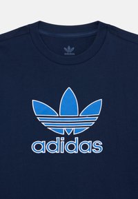 Tumtā zila kokvilnas T-kreklu ar lielu zilu trefoil logotipu un baltu "adidas" tekstu zem logotipa, ar apaļu kakla izgriezumu.