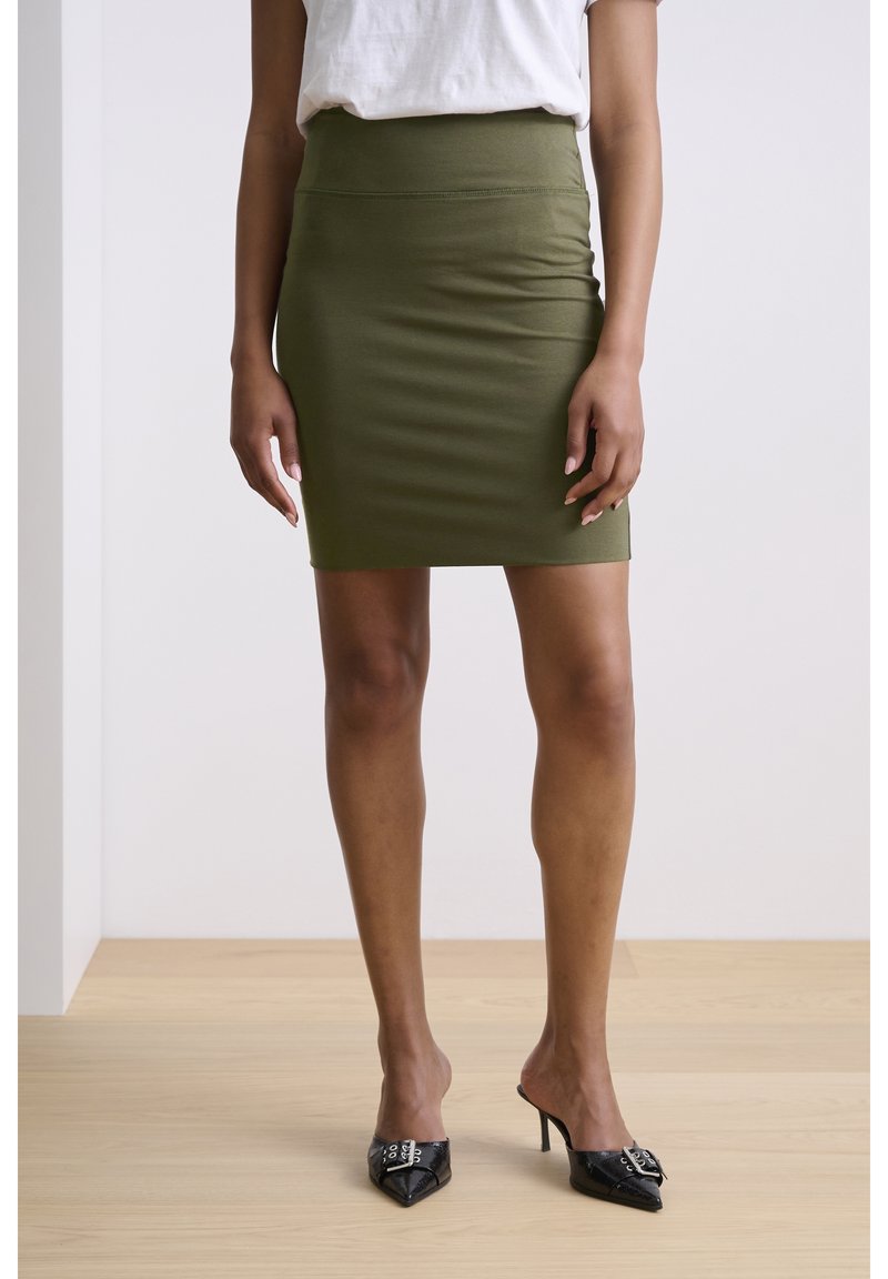 Femme portant un haut blanc, une jupe ajustée vert olive et des mules noires à talons aiguilles, se tenant sur un sol en bois clair.