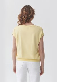 Korte mouwen gele top met een brede halslijn, losse pasvorm en een zachte textuur. Gecombineerd met witte jeans met opgestikte zakken.