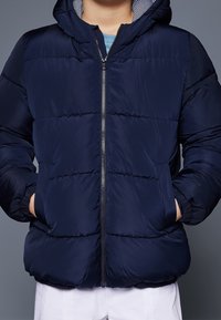 Marineblauwe pufferjack met rits aan de voorkant, capuchon en twee zijzakken. Voorzien van een doorgestikt ontwerp en gestreepte binnenvoering. Gladde stoftextuur.