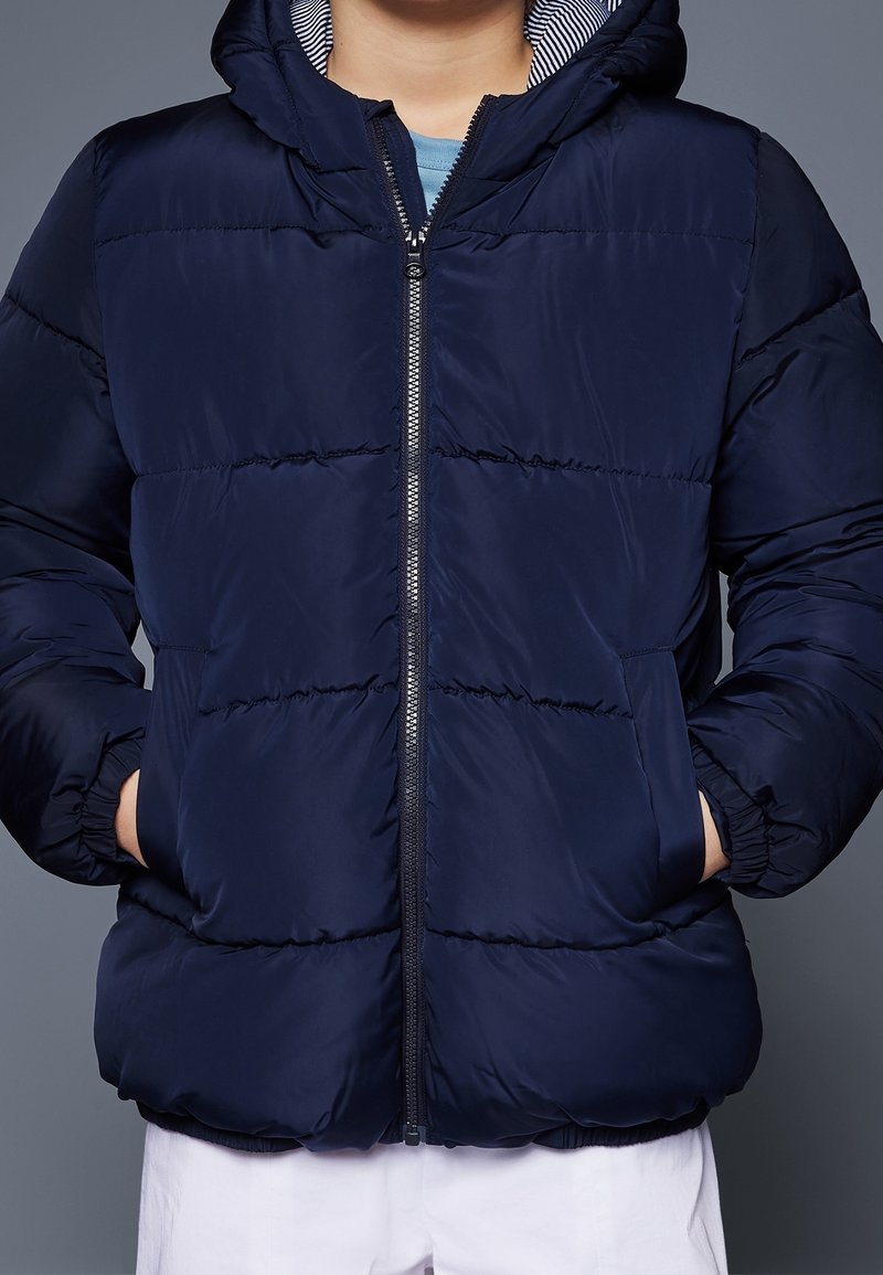 Marineblauwe pufferjack met rits aan de voorkant, capuchon en twee zijzakken. Voorzien van een doorgestikt ontwerp en gestreepte binnenvoering. Gladde stoftextuur.