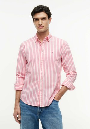 Mężczyzna w różowo-białej koszuli w pionowe paski z guzikami, z podwiniętymi rękawami, pozujący z jedną ręką w kieszeni niebieskich jeansów.