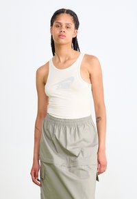 Beige bordás ujjatlan felső, világosszürke Nike logóval, világosszürke, rugalmas derekú szoknyával párosítva, oldalsó zsebekkel.
