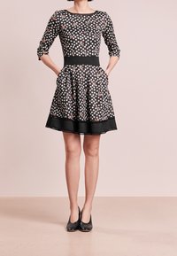 Robe noire à manches courtes, motif floral, taille cintrée, jupe évasée avec poches et bord noir à l'ourlet ; portée avec des talons noirs.