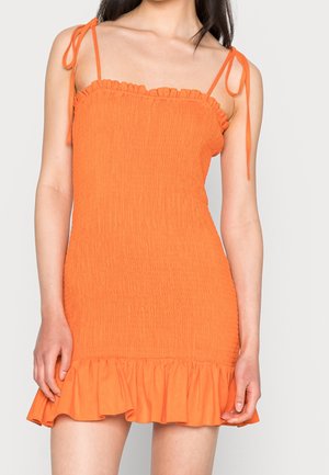 Vrouw draagt een mouwloze oranje mini-jurk met textuur, strikbandjes en een gerimpelde zoom, afgebeeld van schouders tot halverwege de dij.