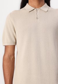 Béžové polo tričko vyrobené z texturovaného pleteného materiálu, s klasickým límcem a knoflíkovou légou na tři knoflíky, s krátkými rukávy.
