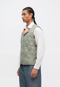 Pieddienēts zaļš bezmugurs veste ar priekšpuses pogu aizdari un kontrastējošu oranzā odere, savienota ar baltu apkakles kreklu un pelēkiem biksēm.