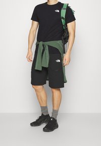 Camiseta negra de manga corta, suéter de punto verde atado a la cintura, pantalones cortos negros con bolsillo, calcetines grises y zapatillas deportivas negras. Detalle de mochila verde.