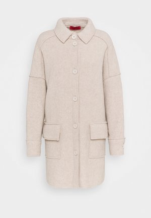 Beige villakangasviitta, jossa on kauluksen muotoinen pääntie, kuusi nappia, kaksi etutaskua ja leveät hihat, joissa on kanttaukset. Sujuva pinta ja räätälöity malli.