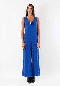 Tuta blu con scollatura a V e un accento metallico circolare sul petto. Realizzata in tessuto morbido, presenta gambe ampie e un corpino aderente.