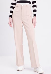 Pantaloni beige su misura con vita alta e pieghe frontali. Tessuto morbido, design a gamba larga. Abbinati a sneakers bianche e una maglietta a righe.
