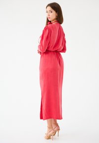 Robe longue en lin de couleur corail vif, avec une taille cintrée, des manches bouffantes et une fente sur le côté. Associée à des escarpins beiges à brides.
