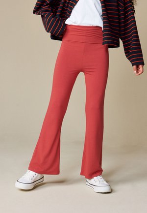 Niño con pantalones rojos de talle alto y campana, zapatillas blancas, camisa blanca y chaqueta azul marino con rayas rojas, de pie sobre un fondo liso.