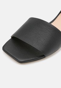 Musta nahkainen liukusandaali, jossa on leveä teksturoitu hihna, neliön muotoinen kärki ja sileä pohja. Minimalistinen muotoilu, ei koristeita tai varusteita.