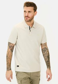 Beige polo med krave, sorte knapdetaljer, korte ærmer og et logo på brystet. Stoffet ser glat ud; afslappet pasform.
