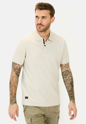 Beige polo med krave, sorte knapdetaljer, korte ærmer og et logo på brystet. Stoffet ser glat ud; afslappet pasform.