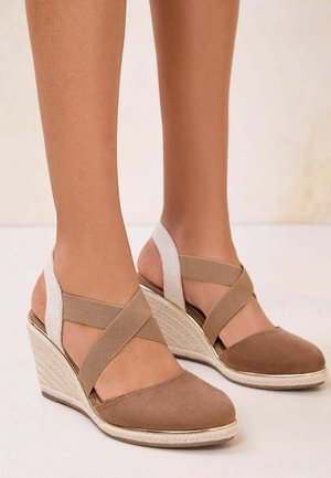 Feet wearing beige wedge espadrilles with crisscross elastic straps and closed toe, standing on a light floor against a neutral background.