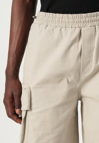 Beige cargo pants with elastic waistband, side pockets, and a textured fabric. Visible stitching detail on the front and side pocket flap.