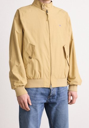 Homme portant une veste jaune moutarde zippée avec des poches à boutons et des poignets côtelés, associée à un jean bleu.