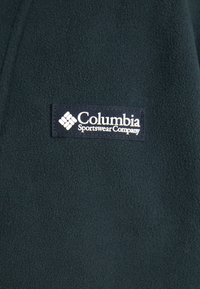 Mörkgrön fleecejacka med en Columbia Sportswear Company-logotyp i vitt på en marinblå bakgrund. Mjuk textur och enkel design.