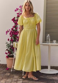 Maxikjole i blød gul, med puffærmer, smocked talje og flagrende nederdel. Stylet med flade sandaler mod en blomsterbaggrund.