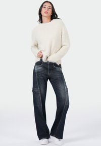 Crème fuzzy trui met lange mouwen, gecombineerd met donkere wijde jeans met voorzakken en witte sneakers.