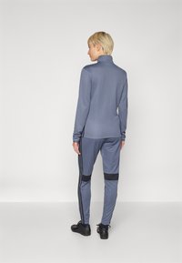 Conjunto atlético gris que incluye una camiseta de manga larga con cuello alto y pantalones a juego con rayas laterales negras y acentos en negro contrastantes en los muslos.