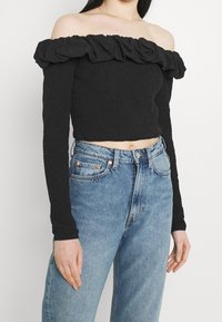 Svart off-shoulder topp med volangkrage, texturerad tyg, kort design, lång ärm, matchad med högmidjade blå jeans.