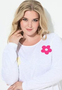 Blondýnka s zelenýma očima, oblečená v bílém topu s dlouhým rukávem zdobeném aplikacemi v růžové a žluté barvě květin, pózující s rukou u obličeje.