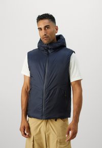 Gilet matelassé bleu marine avec un col haut et une capuche réglable. Il comprend des poches avant zippées et une texture lisse et légère.