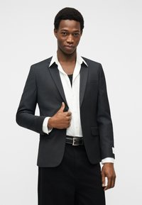 Νέος άνδρας που φοράει μαύρο σακάκι πάνω από άσπρο πουκάμισο, κρατώντας το πέτο του σακακιού με το ένα χέρι, στέκεται μπροστά σε απλό φόντο.