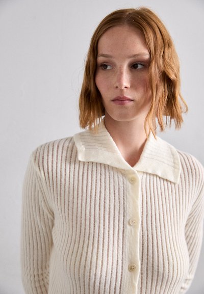 Cardigan crem cu închidere cu nasturi, textură ribbed, dungi contrastante și un guler moale. Materialul pare cald și confortabil.