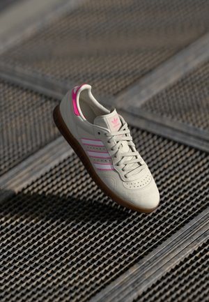 Weißer Adidas-Sneaker mit pinken Streifen und Gummisohle, der auf einer strukturierten Metallgitter-Oberfläche ruht.