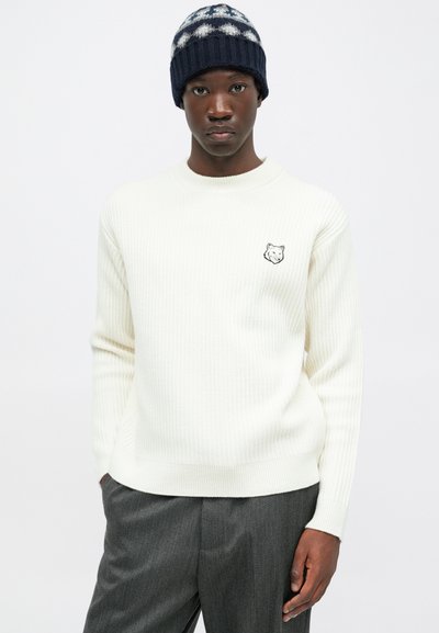 Young man wearing a white ribbed sweater with a small wolf logo, gray trousers, and a blue patterned knit beanie, standing against a white background.