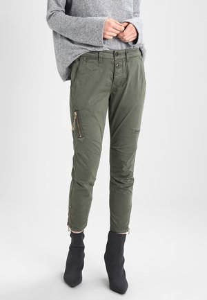 Personne portant un pantalon cargo vert fuselé avec des fermetures éclair sur les poches et les ourlets, associé à des bottines noires et un pull gris.