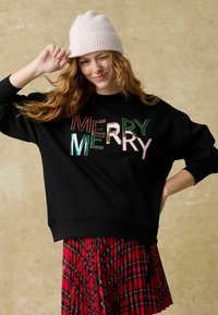 Fekete pulóver, amelyen színes, fényes betűkkel szerepel a "MERRY" felirat. Piros kockás, redőzött szoknyával és világos rózsaszín sapkával párosítva.