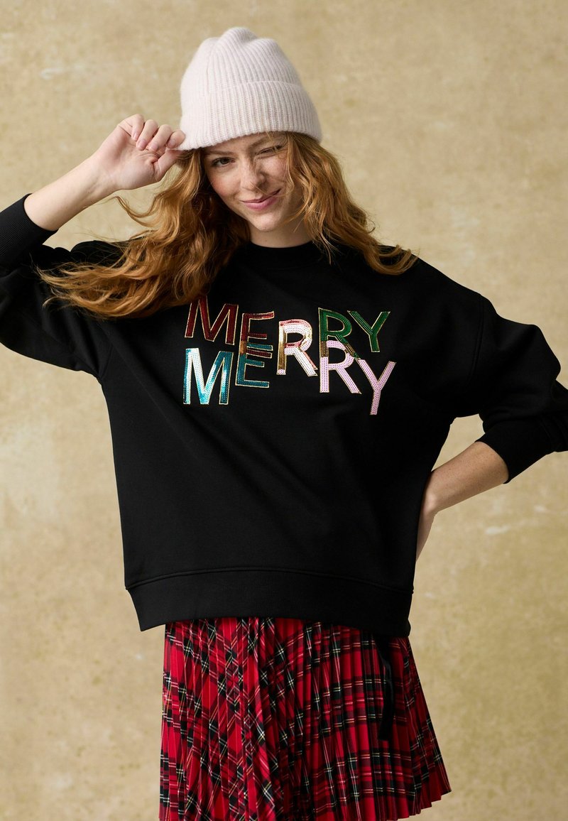 Zwarte sweatshirt met "MERRY" in meerkleurige, glanzende letters. Gecombineerd met een rode geruite plissérok en een lichtroze beanie muts.