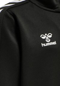 Zwarte sportjacket met een gladde textuur, voorzien van een wit logo en accenten. Inclusief diagonale witte strepen op de schouders.